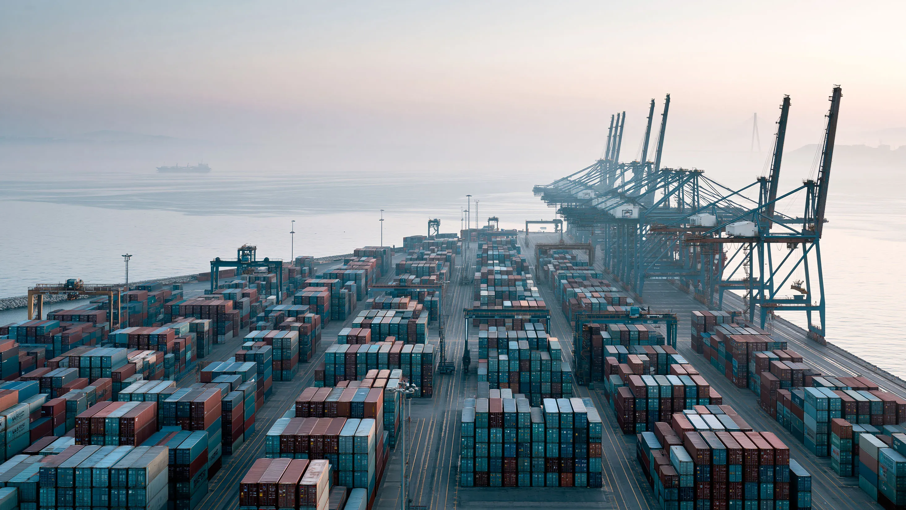 Aerial view of an industrial logistics terminal at dawn, stacked shipping containers and gantry cranes.